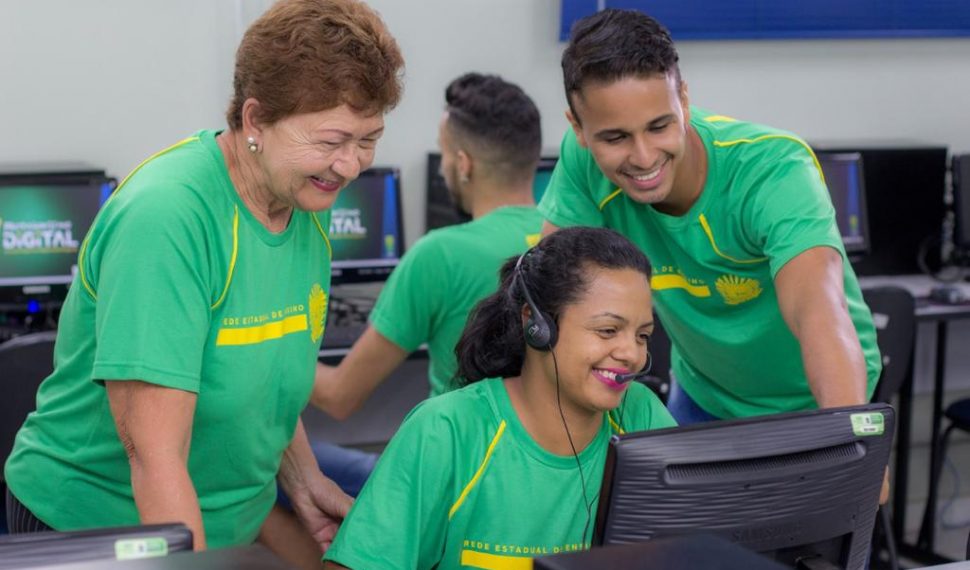 Estudantes da EJA Mato Grosso do Sul estão vestindo camisas verdes atentos à tela do computador. No centro uma pessoa está sentada usando fones de ouvido. e duas estão em pé, uma de cada lado. No fundo uma pessoa está sentada de constas.