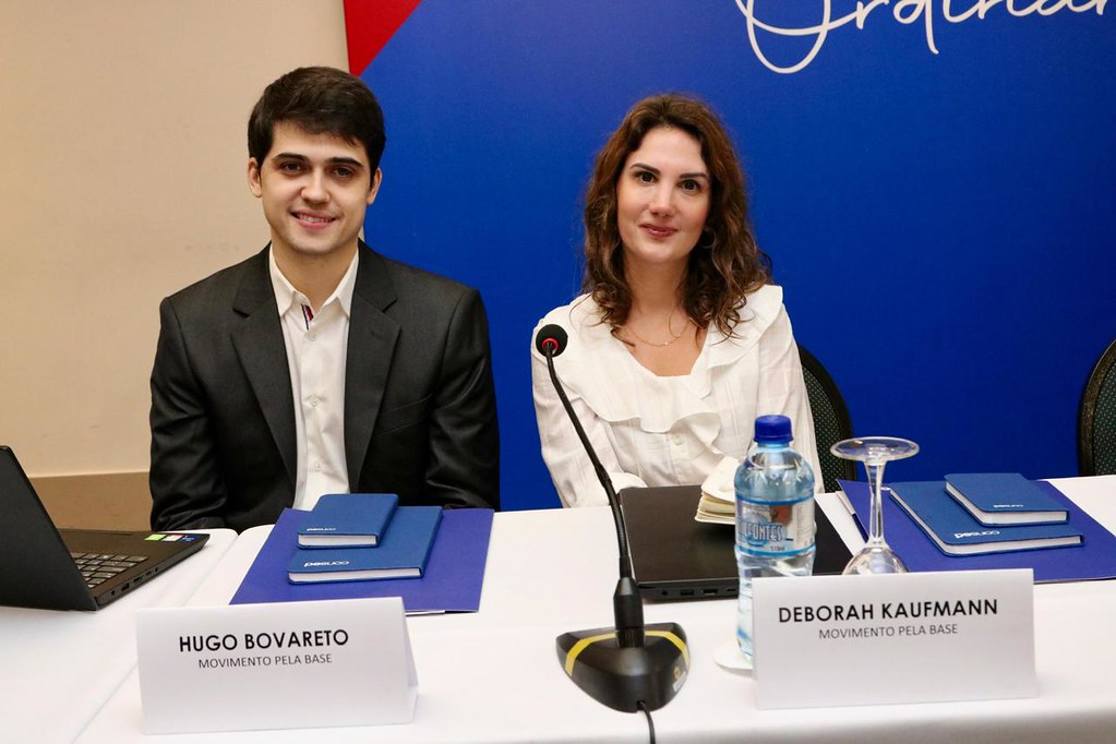 homem branco de cabelo preto usando terno preto e mulher branca de cabelo castanho claro e blusa branca sentados numa mesa de palestra com um microfone de mesa em sua frente